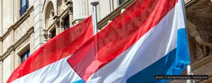 Zwei luxemburgische Flaggen mit roten, weißen und blauen Streifen wehen vor einem historischen Gebäude mit verzierten Fassadenelementen und Säulen im Wind.