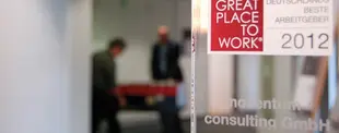 Das Bild zeigt eine Auszeichnung „Great Place to Work 2012“ für noventum consulting GmbH, mit einem unscharfen Hintergrund von Büroangestellten in Bewegung.