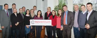 Eine Gruppe von Menschen steht zusammen in einem Raum und hält ein noventum IT MANAGEMENT CONSULTING-Schild,