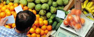 Das Bild zeigt einen Marktstand mit frischem Obst und Gemüse. Ein Händler wiegt eine Tüte mit Orangen auf einer digitalen Waage und notiert die Preise.