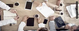 Mehrere Personen sitzen an einem Holztisch mit Notizbüchern, Tablets, Smartphones, Kaffee und Dokumenten und arbeiten gemeinsam an einem geschäftlichen Meeting.