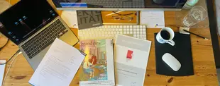Ein Schreibtisch mit Laptop, Tastatur, Monitor, Dokumenten, einer Zeitschrift, einer Brille, Kaffee und Wasser. Darunter eine Brand eins Auszeichnung für Finanzmanagement-Consulting.