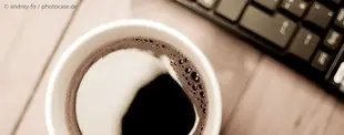 Eine Tasse schwarzer Kaffee auf einem Holztisch, daneben eine Computertastatur – ein symbolisches Bild für Arbeit, Produktivität und digitale Flexibilität.