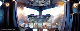 Das Bild zeigt das Cockpit eines Flugzeugs mit zwei Piloten in Bewegung, umgeben von zahlreichen Bedienelementen, Bildschirmen und Instrumenten.