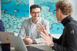 Zwei Männer in Business-Kleidung sitzen an einem Tisch mit Laptops und sprechen gestikulierend. Einer lächelt, während der andere engagiert erzählt.