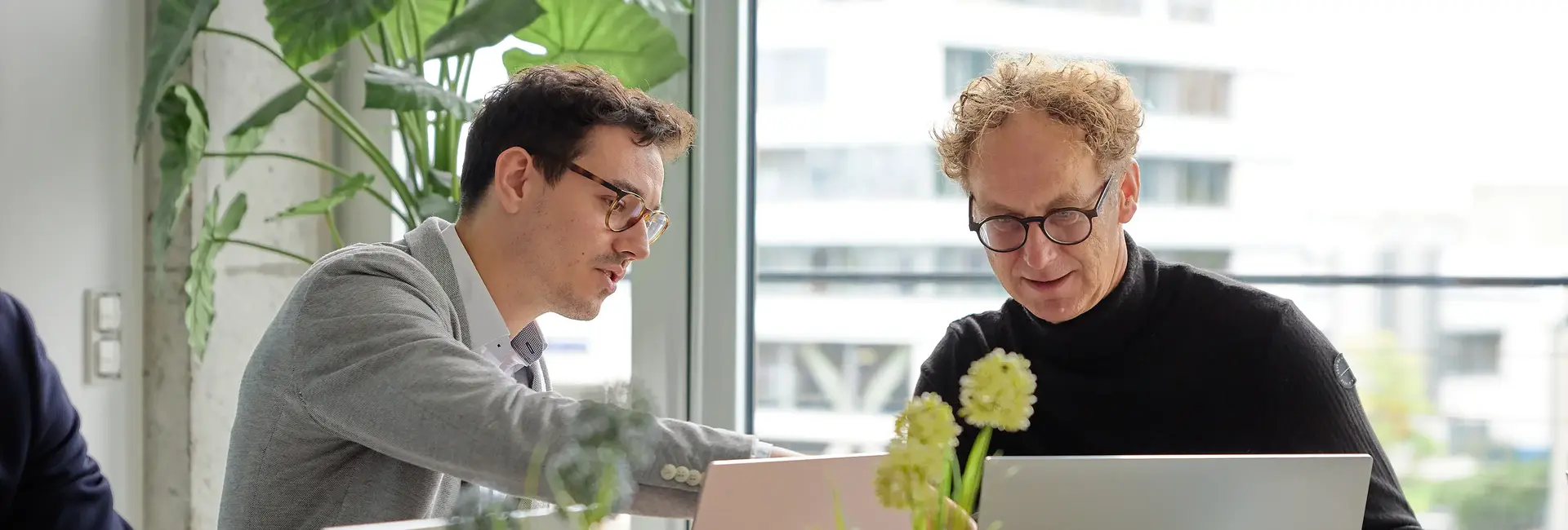 Zwei Männer arbeiten an Laptops, einer zeigt etwas auf dem Bildschirm. Sie sitzen in einem modernen Büro mit großen Fenstern und Pflanzen im Hintergrund.