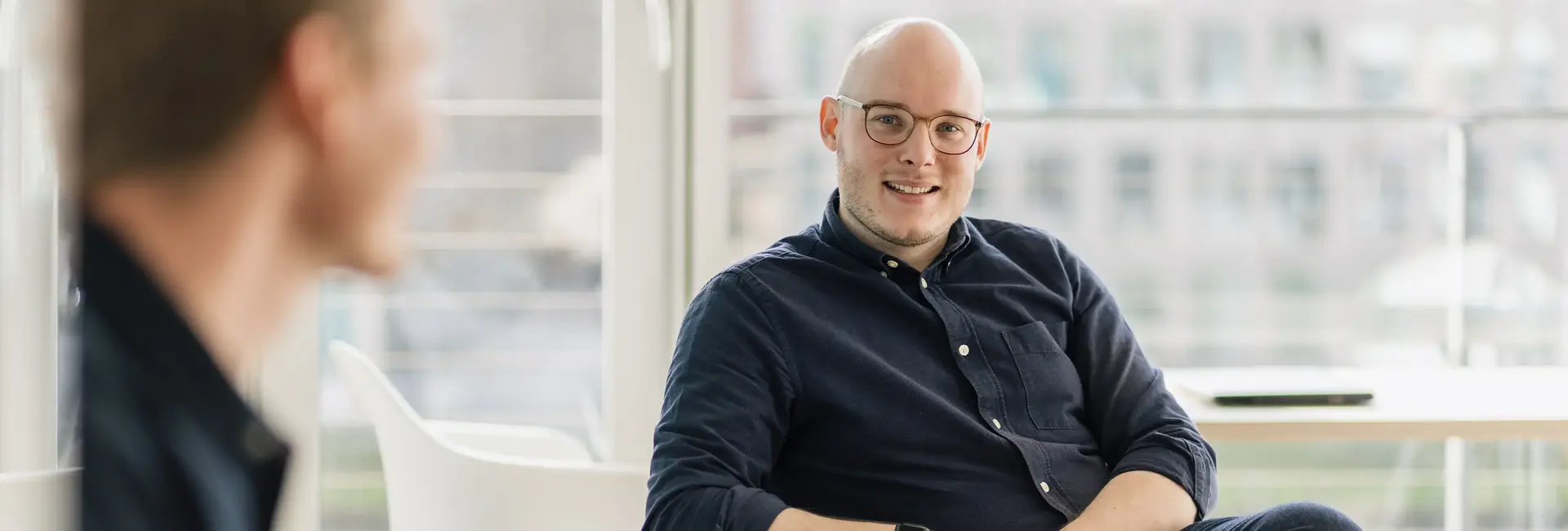 Marc Schwellenbach sitzt und lächelt in einem modernen, lichtdurchfluteten Büro. Im Vordergrund ist eine weitere Person leicht unscharf.