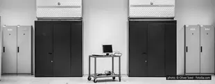 Ein schwarz-weißes Foto eines Serverraums mit zwei großen Serverschränken, Schaltschränken an den Seiten und einem mobilen Wagen mit Monitor und Kabeln in der Mitte.