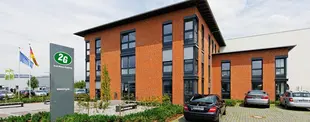Ein modernes Bürogebäude mit roter Klinkerfassade, großen Fenstern und 2G-Logo. Im Vordergrund Parkplätze, ein Firmenschild und wehende Flaggen.