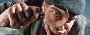 Ein Mann mit Mütze betrachtet etwas durch eine Lupe, sein Auge wird dabei stark vergrößert. Das Bild symbolisiert Untersuchung, Analyse oder Detailgenauigkeit.