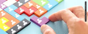 Bunte Puzzleteile mit weißen Personensymbolen werden zusammengesetzt. Eine Hand fügt ein letztes Teil ein.