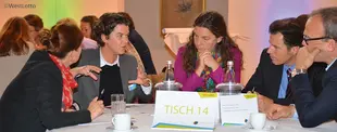 Mehrere Personen diskutieren engagiert an Tisch 14 während eines Workshops, umgeben von Notizen, Wasser und Kaffeetassen.