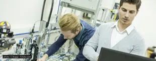 Zwei Ingenieure arbeiten in einem Labor: Einer bedient ein Laptop, der andere justiert eine Maschine mit technischen Komponenten und Kabeln im Hintergrund.