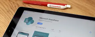 Ein Tablet zeigt die Microsoft SharePoint-App im App Store. Daneben liegt ein noventum-Kugelschreiber auf einem Holztisch.
