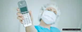 Eine Person in OP-Kleidung mit Maske und Haube hält ein schnurloses Telefon in die Kamera, der Fokus liegt auf dem Telefon, der Hintergrund ist unscharf.