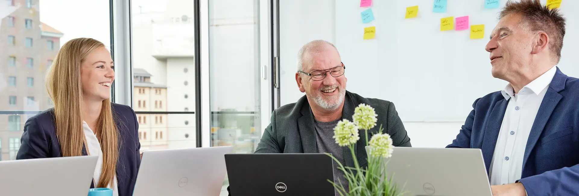 Drei Personen sitzen in einem modernen Büro mit Laptops und Whiteboard im Hintergrund.