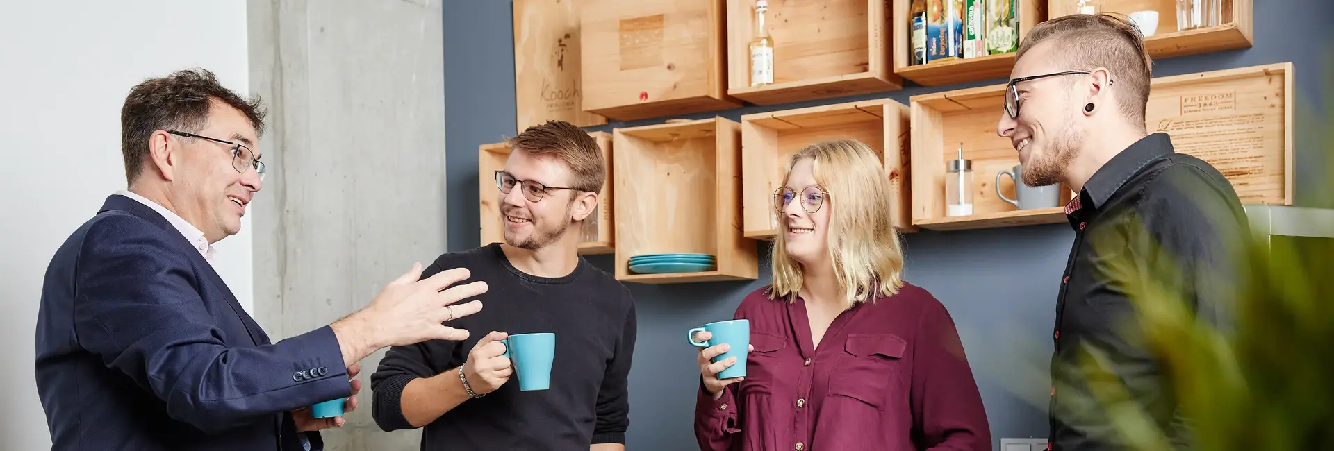 Vier Personen unterhalten sich entspannt in einer Büroküche. Sie halten Tassen in der Hand und lachen gemeinsam während der Kaffeepause.