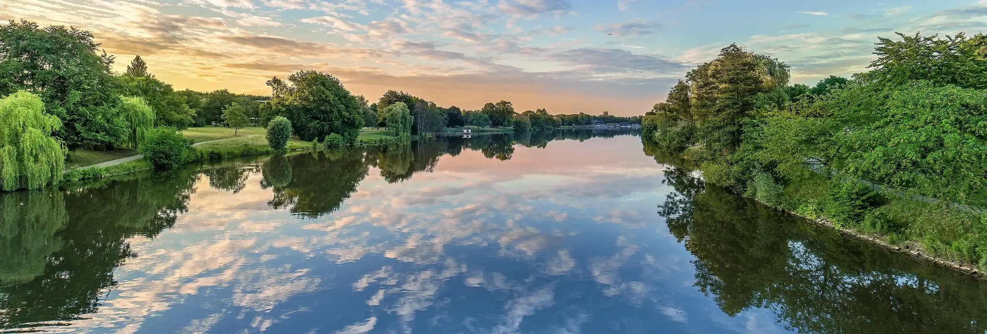 Der Aasee in Münster spiegelt den Himmel bei Sonnenuntergang und ist umgeben von Bäumen und Wiesen.