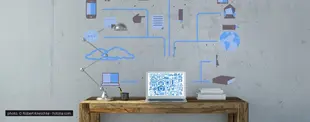 Ein Laptop auf einem Holztisch vor einer Betonwand mit digitalen Netzwerksymbolen, die Cloud, Kommunikation und globale Vernetzung darstellen.