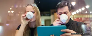 Ein Mann und eine Frau trinken Kaffee in einem Café. Der Mann hält ein Tablet, während sie gemeinsam auf den Bildschirm schauen. Warmes Licht im Hintergrund.