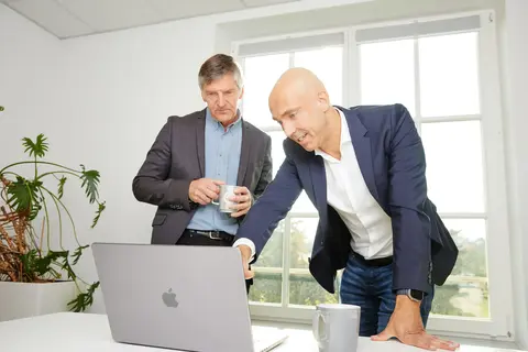Zwei Männer in Business-Kleidung stehen an einem Tisch mit Laptop und Kaffeetassen. Einer lehnt sich vor und zeigt auf den Bildschirm, während der andere zuhört.