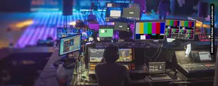 Ein Techniker sitzt an einem Mischpult mit vielen Monitoren und Steuergeräten in einer Eventhalle. Die Bildschirme zeigen Testbilder und Live-Feeds der Veranstaltung.