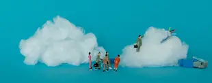 Miniaturfiguren stehen vor Wolken aus Watte, die mit Netzwerkkabeln verbunden sind. Eine Figur erklimmt eine Wolke, symbolisiert Cloud-Technologie und Vernetzung.