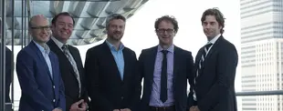 Fünf Männer in Anzügen stehen auf einer Terrasse mit moderner Glasfassade, lächeln in die Kamera. Im Hintergrund sind hohe Bürogebäude zu sehen.