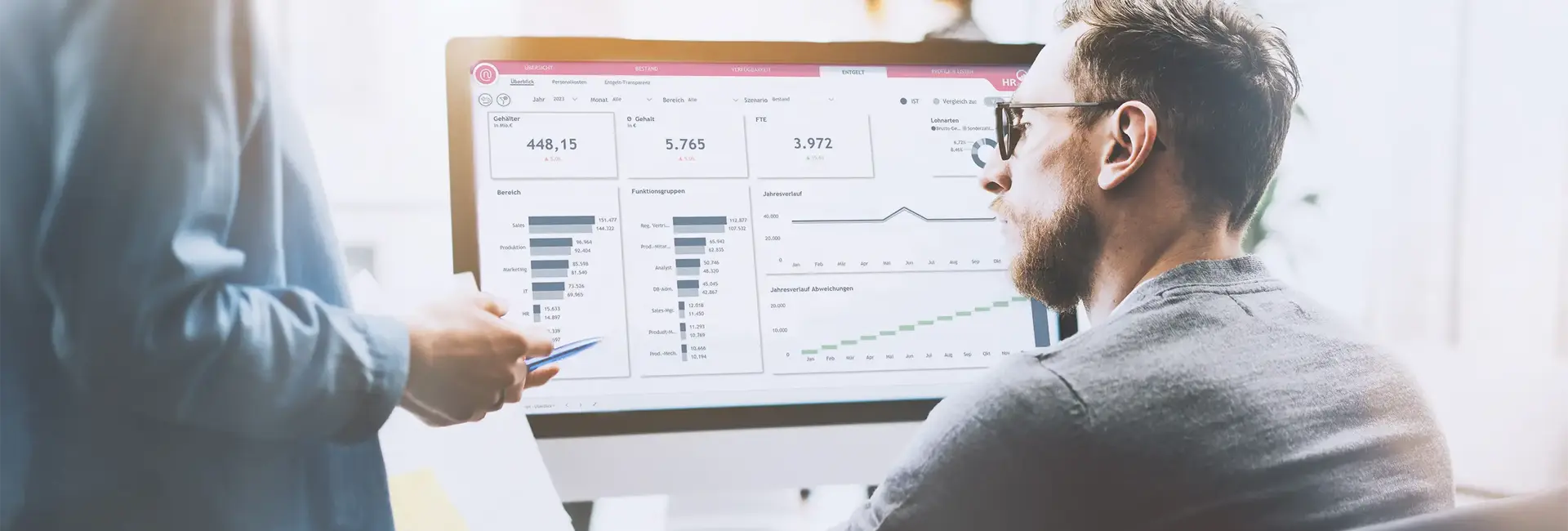 Ein Mann schaut konzentriert auf einen Bildschirm mit HR-Analytics-Daten, während eine Person daneben mit einem Stift auf die Visualisierungen zeigt.