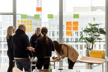Ein kreativer Workshop mit mehreren Personen, die an einem Tisch arbeiten, Ideen aufschreiben und an Fenstern mit Post-its eine Ideensammlung gestalten.