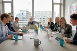 Eine Gruppe von Geschäftsleuten sitzt in einem modernen Büro an einem großen Tisch, arbeitet mit Laptops und diskutiert konzentriert. Helle Fensterfront im Hintergrund.