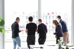 Fünf Personen stehen vor einer großen Fensterfront und arbeiten mit Klebezetteln an der Scheibe. Sie diskutieren und sammeln Ideen in einer kreativen Workshop-Atmosphäre.