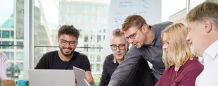 Ein Team arbeitet gemeinsam an Laptops in einem modernen Büro mit großen Fenstern. Eine Person erklärt etwas, während die anderen interessiert zuhören und lächeln.