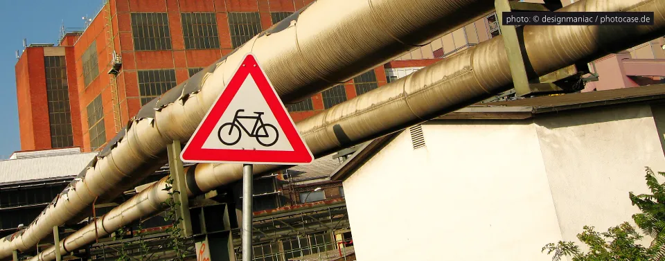 Ein Verkehrsschild mit einem Fahrradsymbol vor einer Industriekulisse mit großen Rohrleitungen und Backsteingebäuden unter einem sonnigen Himmel.