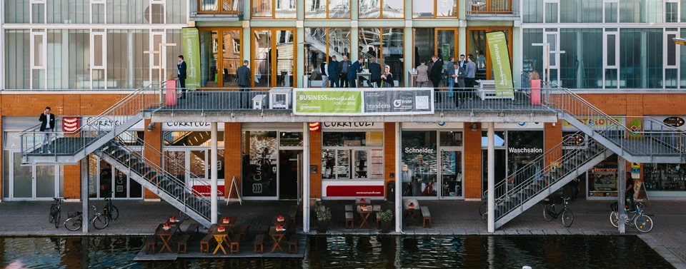 Ein modernes Gebäude mit einer Terrasse über dem Wasser, auf der Menschen bei einer Business-Veranstaltung stehen. Unten befinden sich Geschäfte und ein Café mit Sitzplätzen.