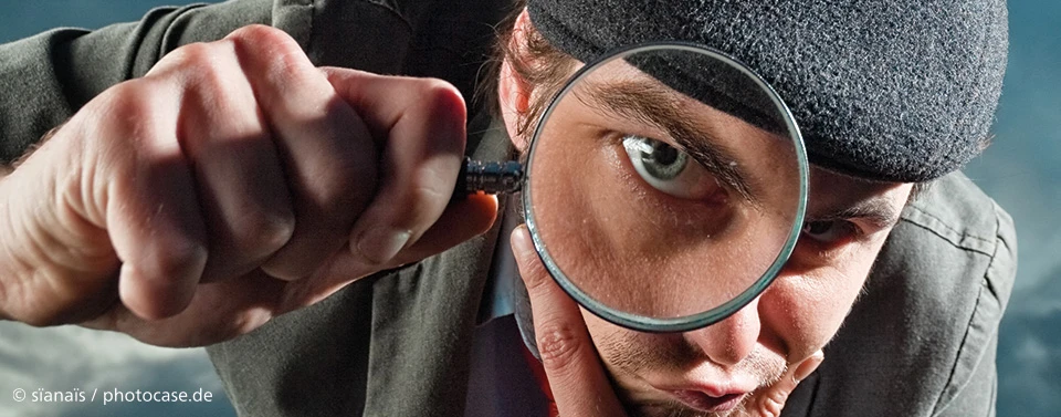 Ein Mann mit Mütze betrachtet etwas durch eine Lupe, sein Auge wird dabei stark vergrößert. Das Bild symbolisiert Untersuchung, Analyse oder Detailgenauigkeit.
