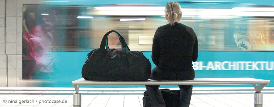 ChatGPT:  Eine Frau sitzt auf einer Bank in einer U-Bahn-Station mit einer schwarzen Tasche neben sich, während eine blaue Bahn mit Bewegungsunschärfe vorbeifährt