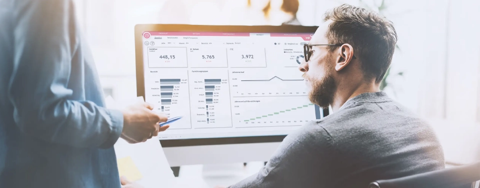 Ein Mann sitzt vor einem Monitor mit HR-Analysedaten, während eine Person daneben mit Stift und Notizblock steht. Helles Büro mit natürlichem Licht im Hintergrund.