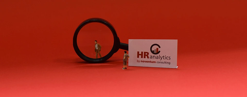 Eine Lupe vergrößert eine Miniaturfigur vor rotem Hintergrund. Daneben steht eine Karte mit dem Logo „HR Analytics by noventum consulting“.
