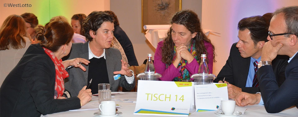 Mehrere Personen diskutieren engagiert an Tisch 14 während eines Workshops, umgeben von Notizen, Wasser und Kaffeetassen.