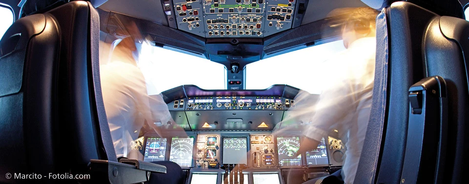 Das Bild zeigt das Cockpit eines Flugzeugs mit zwei Piloten in Bewegung, umgeben von zahlreichen Bedienelementen, Bildschirmen und Instrumenten.