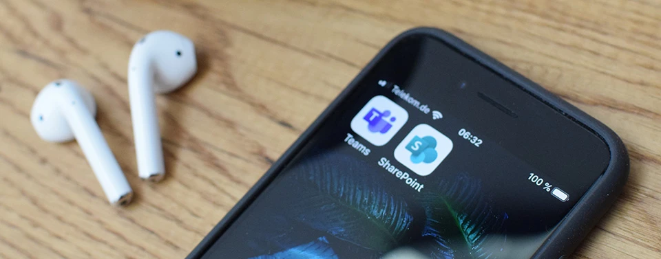 Ein Smartphone mit den Apps Microsoft Teams und SharePoint liegt auf einem Holztisch. Daneben befinden sich zwei kabellose weiße Ohrhörer.