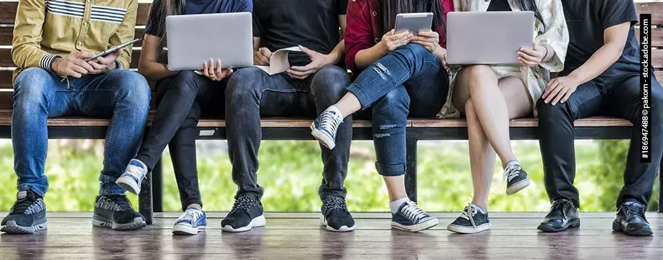 Mehrere Personen sitzen auf einer Bank und nutzen Tablets, Laptops und Smartphones, während sie sich auf ihre digitalen Geräte konzentrieren.
