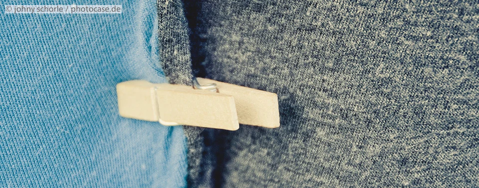 Das Bild zeigt eine hölzerne Wäscheklammer, die zwei unterschiedlich farbige Stoffe, einen blauen und einen grauen, zusammenhält. Nahaufnahme mit Texturdetails.
