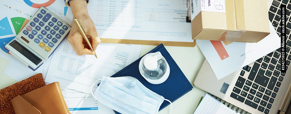 Ein Schreibtisch mit Laptop, Dokumenten, Taschenrechner, Paket, Maske und Desinfektionsmittel und eine schreibende Hand.