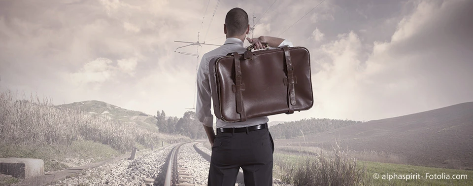Ein Mann im Anzug mit einer Ledertasche steht auf Bahngleisen und blickt in die Ferne, symbolisierend Aufbruch, Veränderung oder eine ungewisse Zukunft