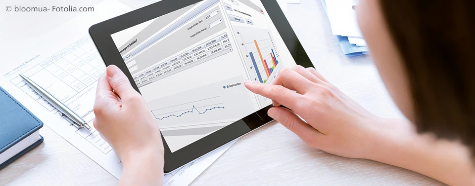 Eine Person nutzt ein Tablet zur Analyse von Geschäftsdaten mit Diagrammen und Tabellen, während auf dem Schreibtisch Unterlagen und ein Stift liegen.