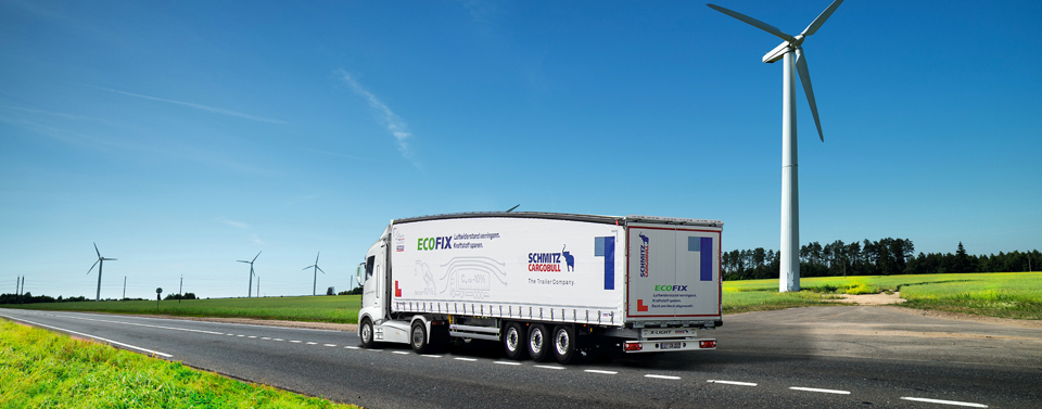 Ein Lkw mit Schmitz Cargobull Anhänger fährt auf einer Landstraße, umgeben von grünen Feldern und Windrädern.