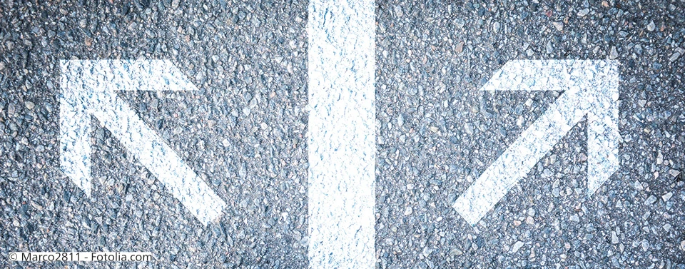 Zwei weiße Pfeile auf Asphalt zeigen in entgegengesetzte Richtungen, getrennt durch eine Mittellinie, symbolisch für Entscheidungen oder verschiedene Wege.