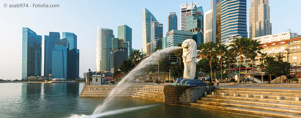 Das Bild zeigt die Skyline von Singapur mit dem berühmten Merlion-Brunnen im Vordergrund, der Wasser in die Bucht speit, umgeben von modernen Hochhäusern.